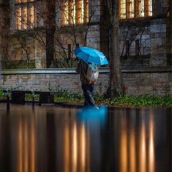 Person with blue umbrella walks at night on campus of Yale University. Illuminated windows on background reflected on water in foreground.